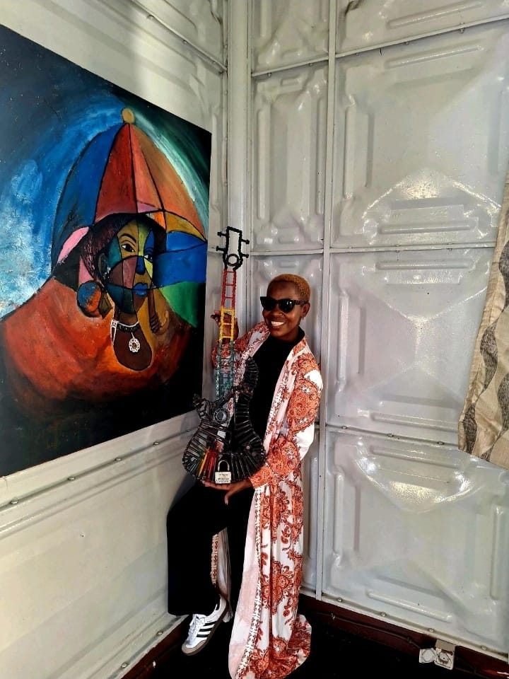 Eunice holding a guitar made of recycled parts beside a painting inside the Iron House in Maputo